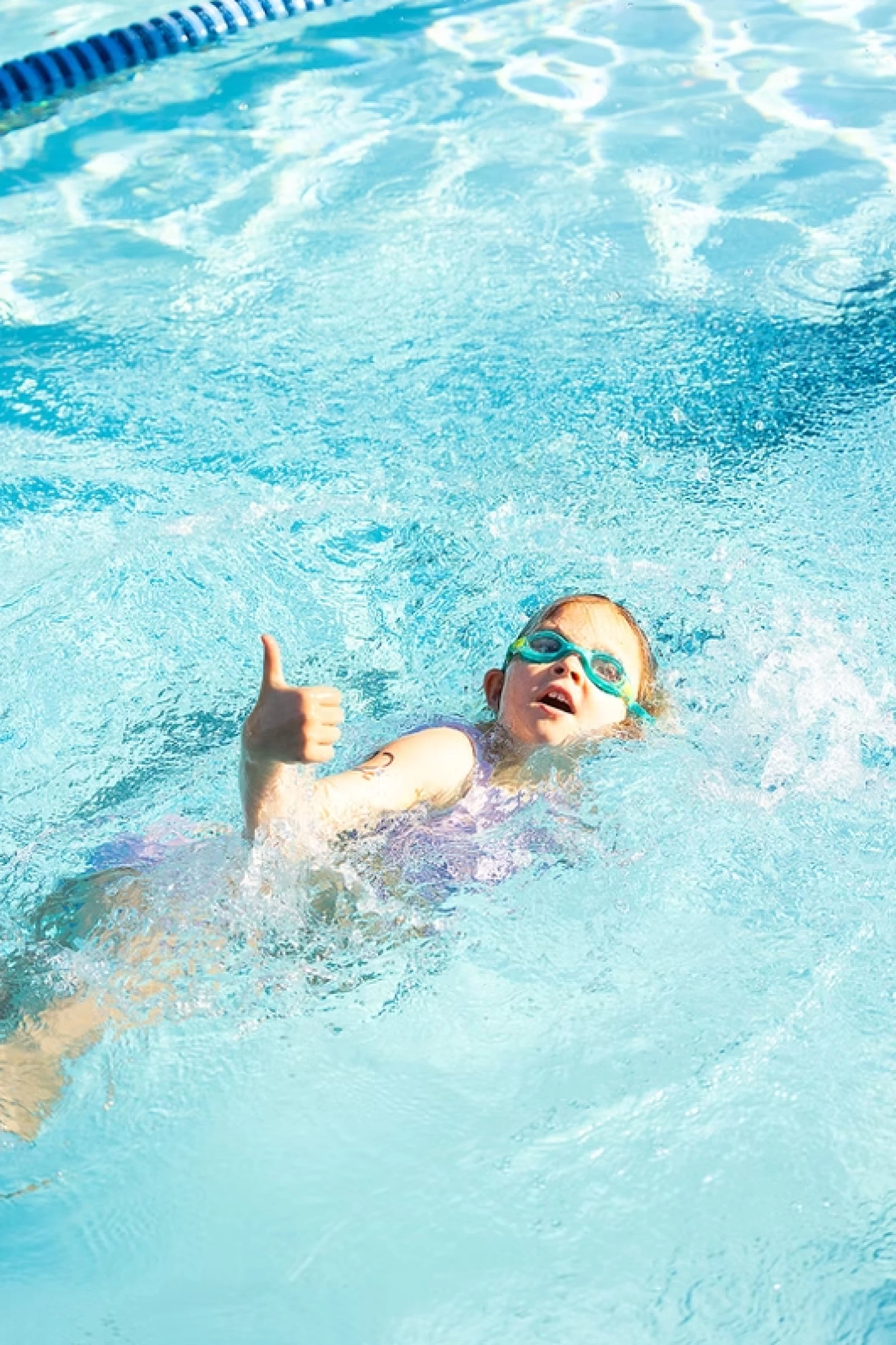 A kid wearing swimming goggles gives a thumbs-up while floating and splashing in a bright blue pool, enjoying a sunny day at swim time.