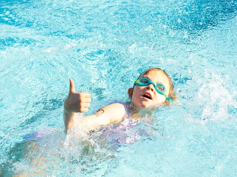 A kid wearing swimming goggles gives a thumbs-up while floating and splashing in a bright blue pool, enjoying a sunny day at swim time.