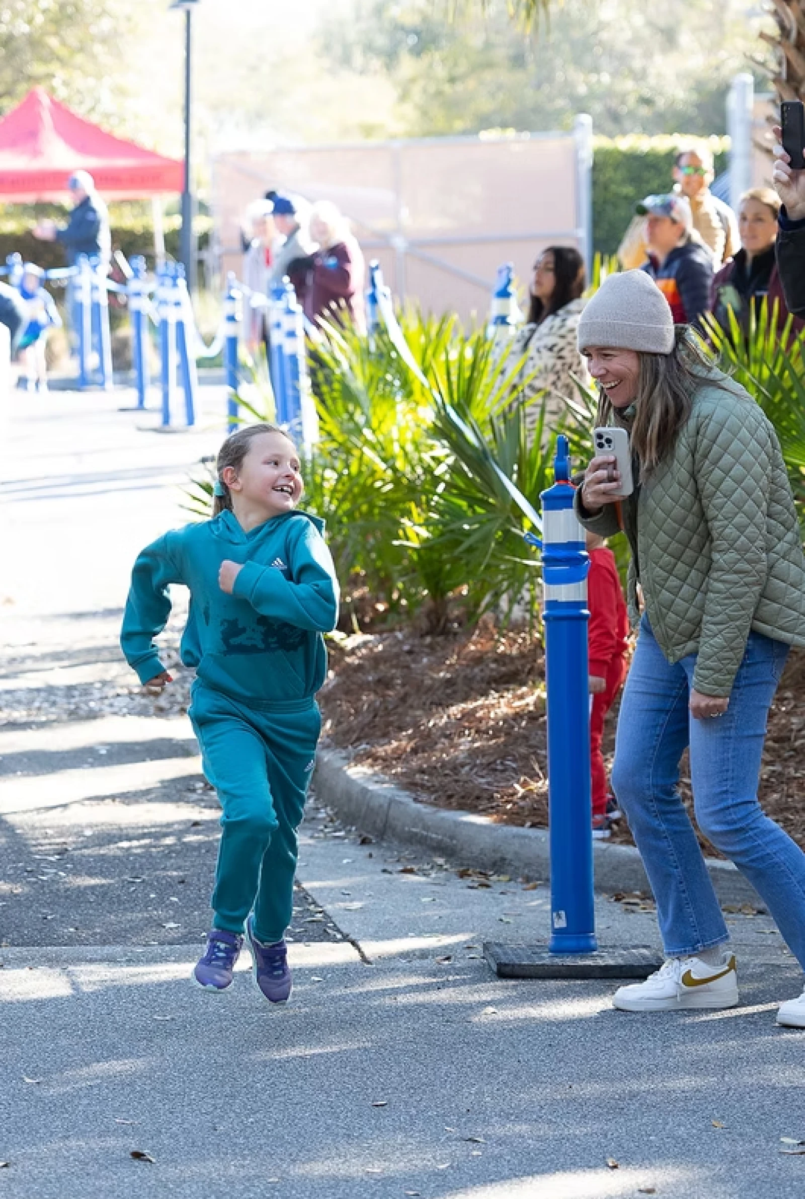 A child in teal runs joyfully toward a laughing woman holding a drink; a sunny outdoor event with tents and onlookers in the background.