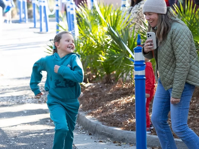 A child in teal runs joyfully toward a laughing woman holding a drink; a sunny outdoor event with tents and onlookers in the background.