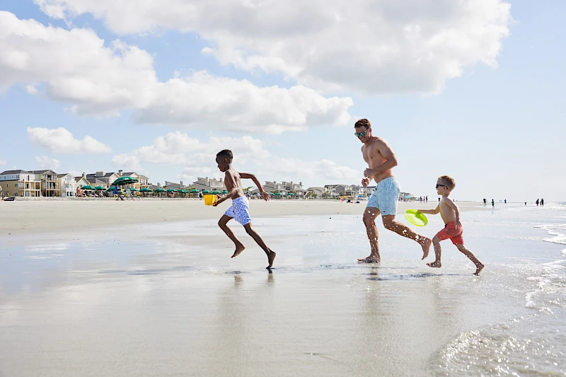 Three kids with a man run along a sunny beach, splashing near the shallow waves, with houses and umbrellas in the background.