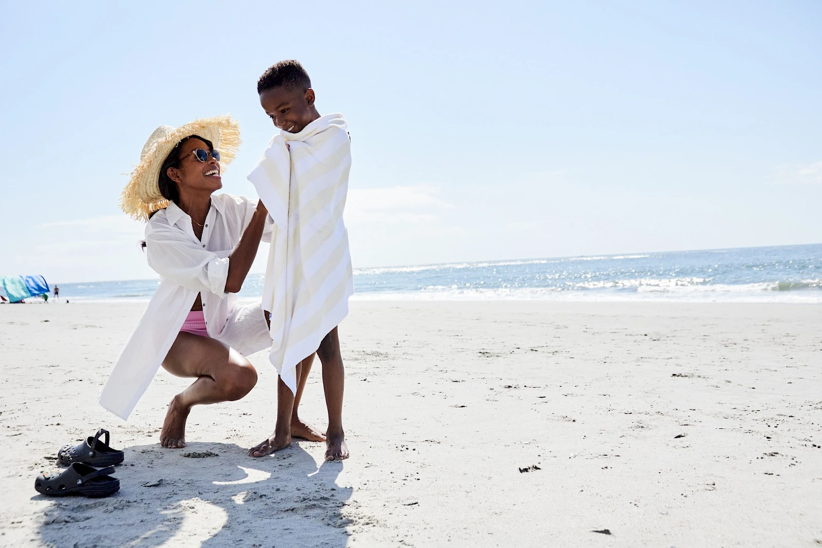 Two people in white beach attire share a moment on the sand by the sea, one kneeling and proposing or adjusting the towel, with sandals nearby.
