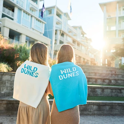 Two people with capes reading “WILD DUNES” walking outside a modern building, sunlight behind them, steps and greenery around. End.