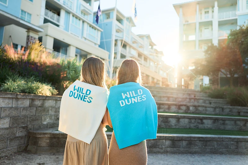 Two people with capes reading “WILD DUNES” walking outside a modern building, sunlight behind them, steps and greenery around. End.