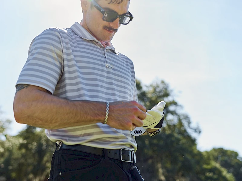 A man in a striped polo and sunglasses checks a pocket-sized device outdoors, with the sun bright behind him and trees in the background.