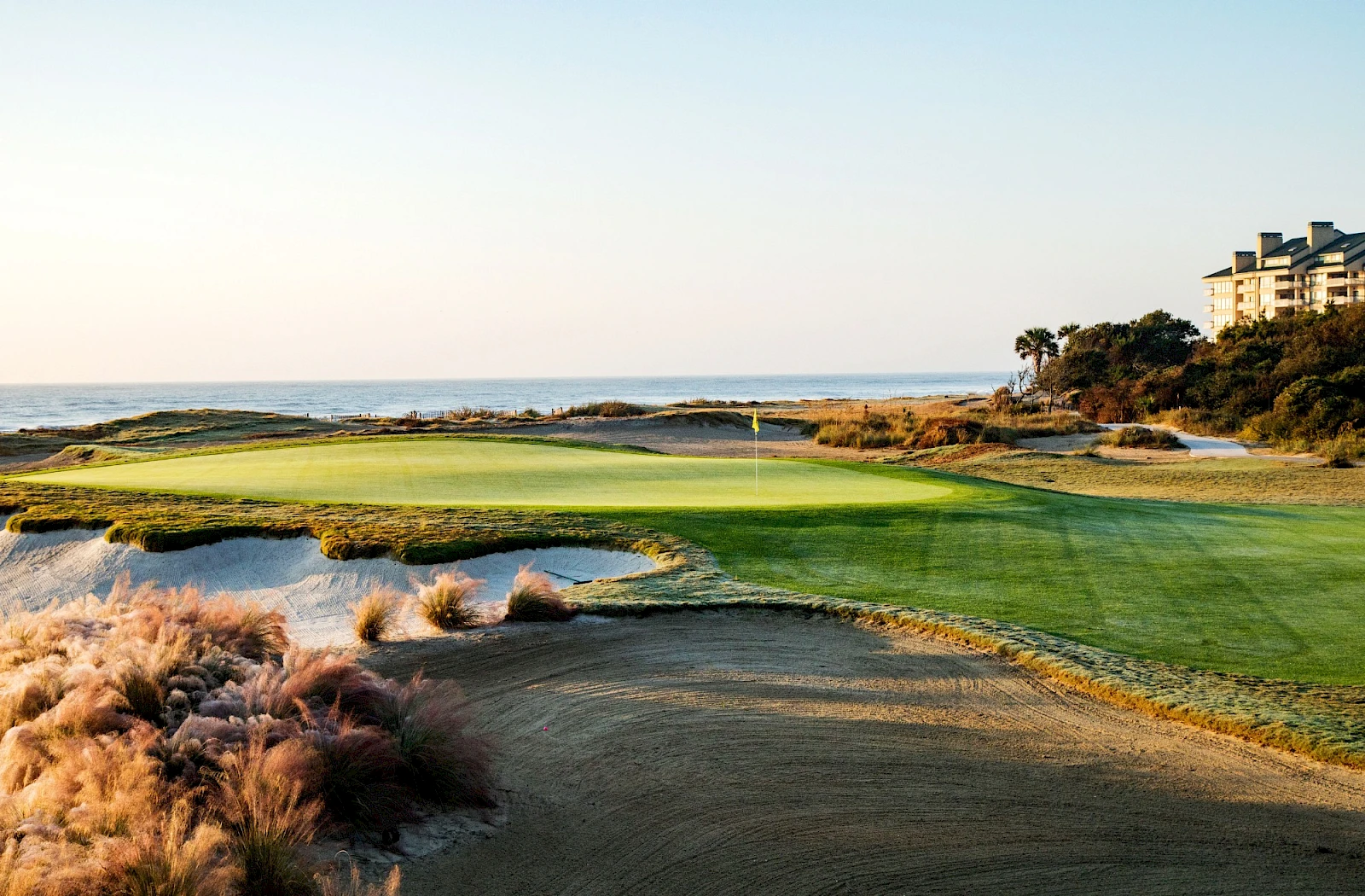 A coastal golf course with green fairways, a sandy bunker, and waves crashing near the rough, under a clear sky at dusk.