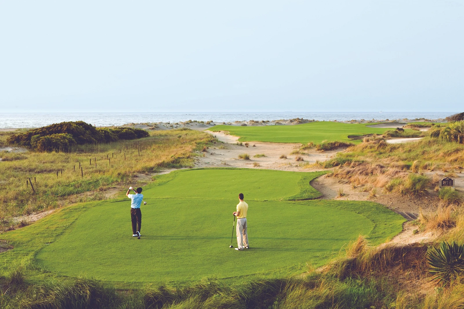 Two golfers stand on a green tee box overlooking a coastal course with rough, sand dunes, and the sea in the distance.