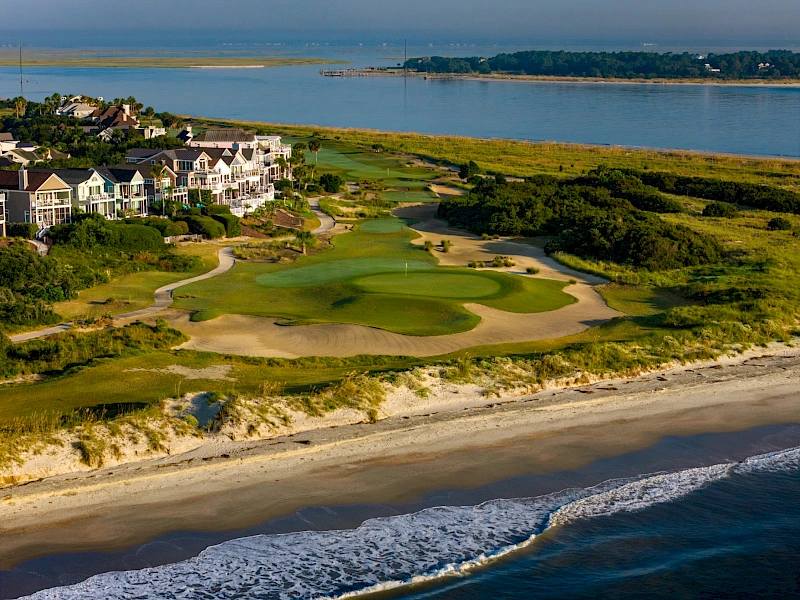 Coastal golf course with green fairways, sandy bunkers, sea wall, and beachfront houses along the shore, blue ocean meeting the horizon.