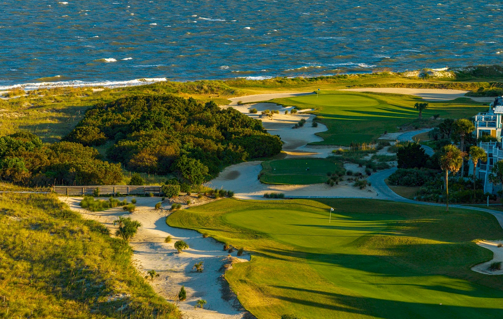 A coastal golf course with green fairways winding between sand dunes and shrubs under a bright sky, with water in the distance.