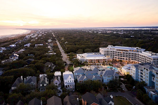 Coastal neighborhood at dusk with a mix of houses and a large curved hotel/condo building near the shore, calm streets, and a sandy horizon.