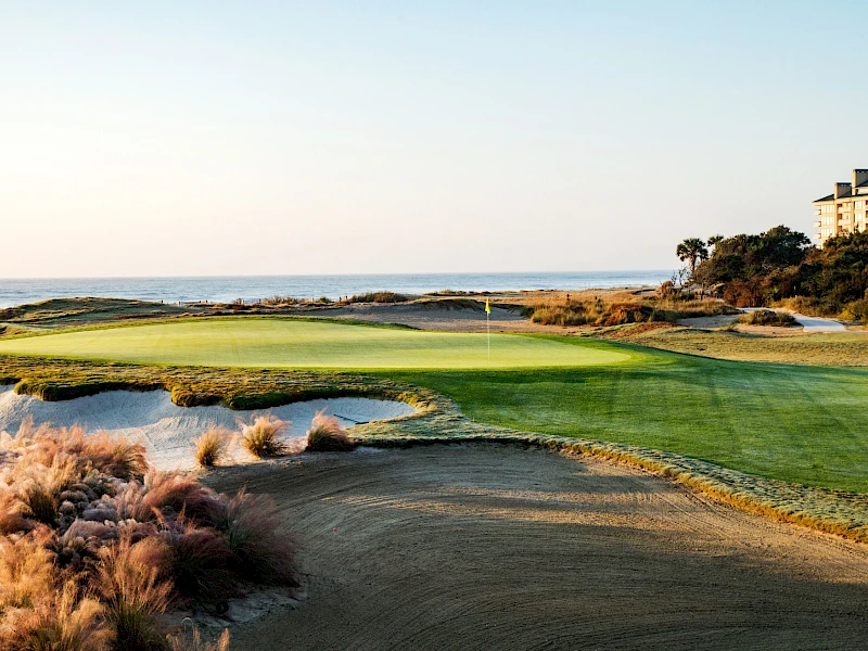 A coastal golf course with green fairways, rough brush, and a sandy hazard near the sea under a clear sky.