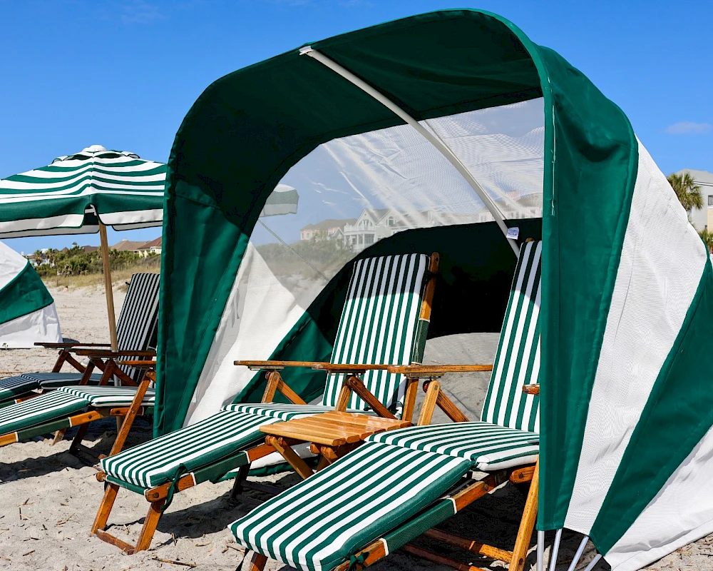 A sunny beach scene with striped lounge chairs under a green-and-white canopy, shaded by beach umbrellas along the sandy shore.