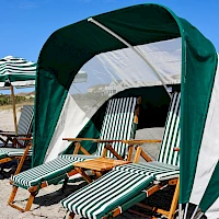 A sunny beach scene with striped lounge chairs under a green-and-white canopy, shaded by beach umbrellas along the sandy shore.