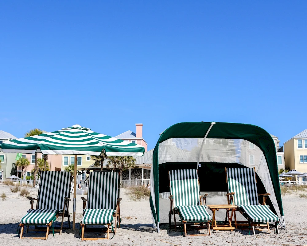 Beach scene with striped lounge chairs, umbrellas, and a shaded cabana on sandy shore under a clear blue sky.
