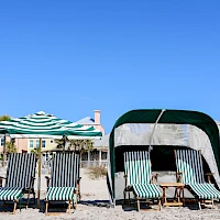 Beach scene with striped lounge chairs, umbrellas, and a shaded cabana on sandy shore under a clear blue sky.