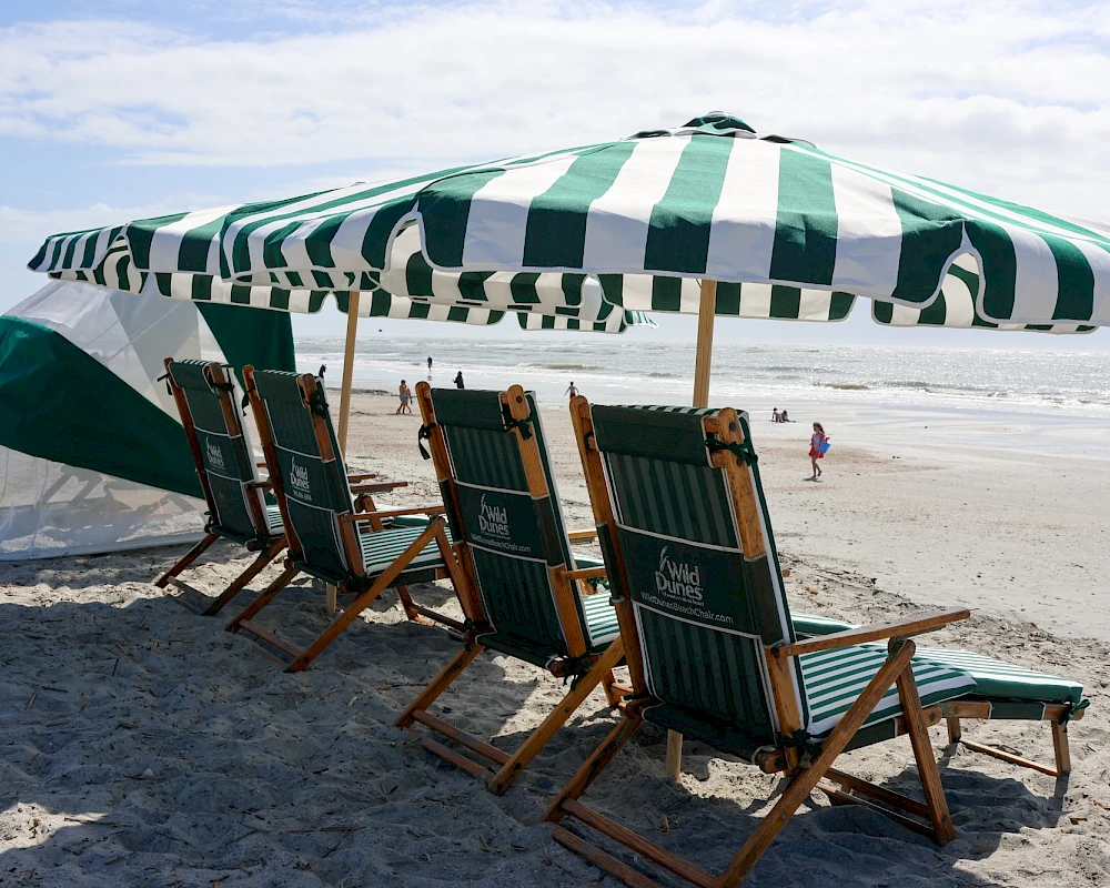People relaxing on a sunny beach under striped umbrellas with wooden lounge chairs, ocean waves in the background, and a bright sky.
