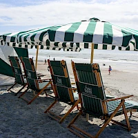 People relaxing on a sunny beach under striped umbrellas with wooden lounge chairs, ocean waves in the background, and a bright sky.