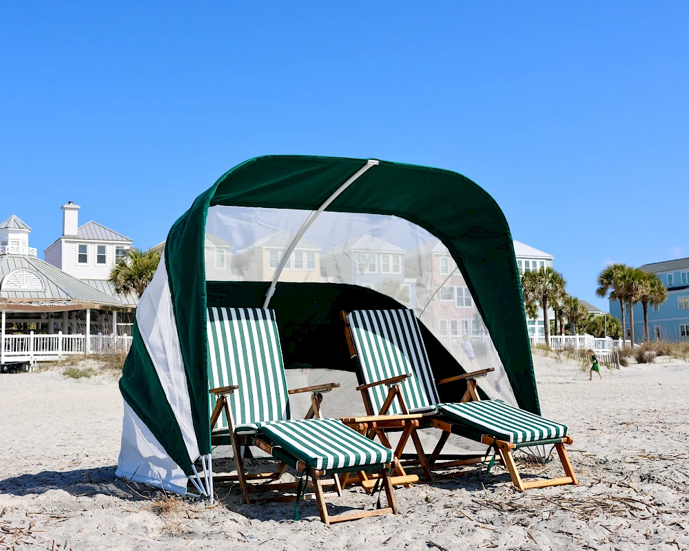 Beach scene with a green and white striped cabana and four lounge chairs on sandy shores, clear blue sky, coastal houses in the background.