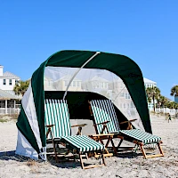 Beach scene with a green and white striped cabana and four lounge chairs on sandy shores, clear blue sky, coastal houses in the background.