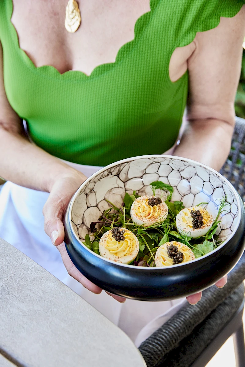 A person in a green top holds a bowl with deviled eggs garnished with roe or seeds. 140 chars ending with a period.