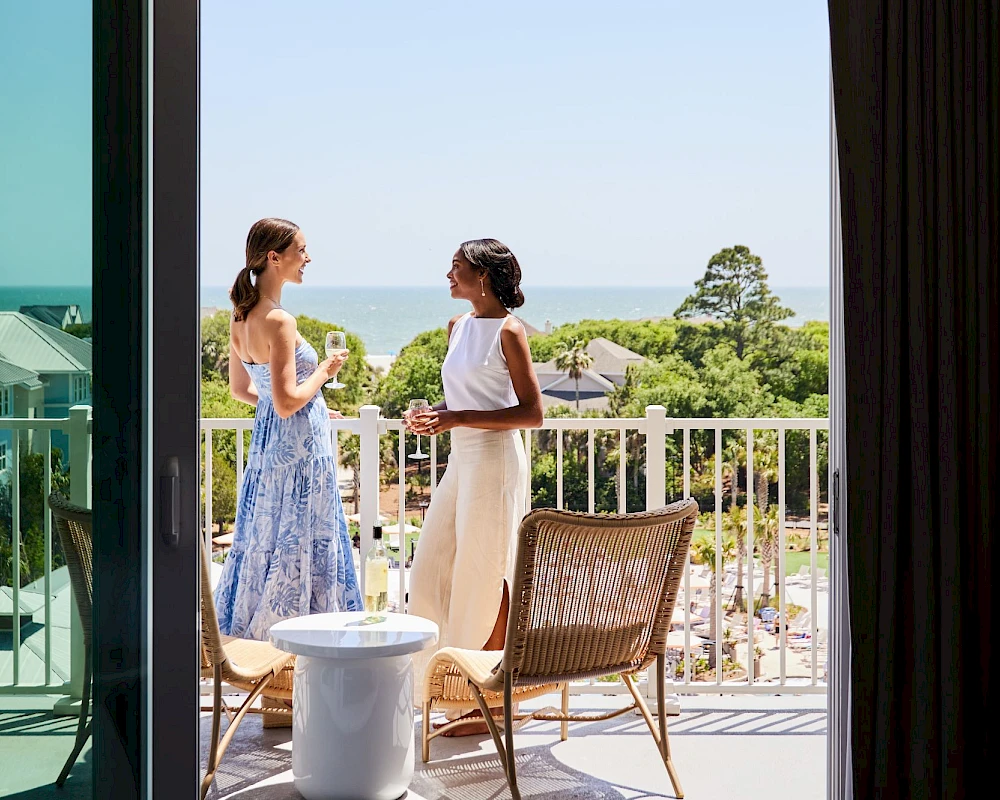 Two women stand on a balcony talking, one in a light dress and the other in a white top and skirt, with chairs and a small table nearby.