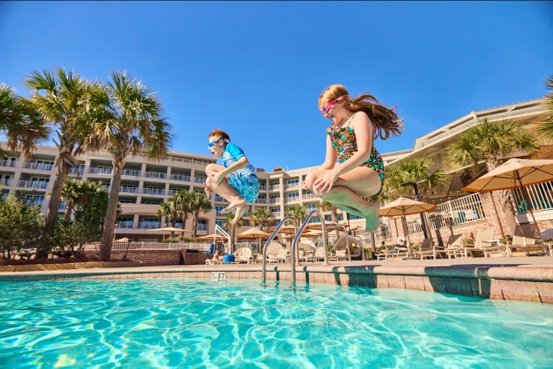 Two kids are jumping into a sunny hotel pool, mid-air, with palm trees and resort buildings in the background, umbrellas nearby.