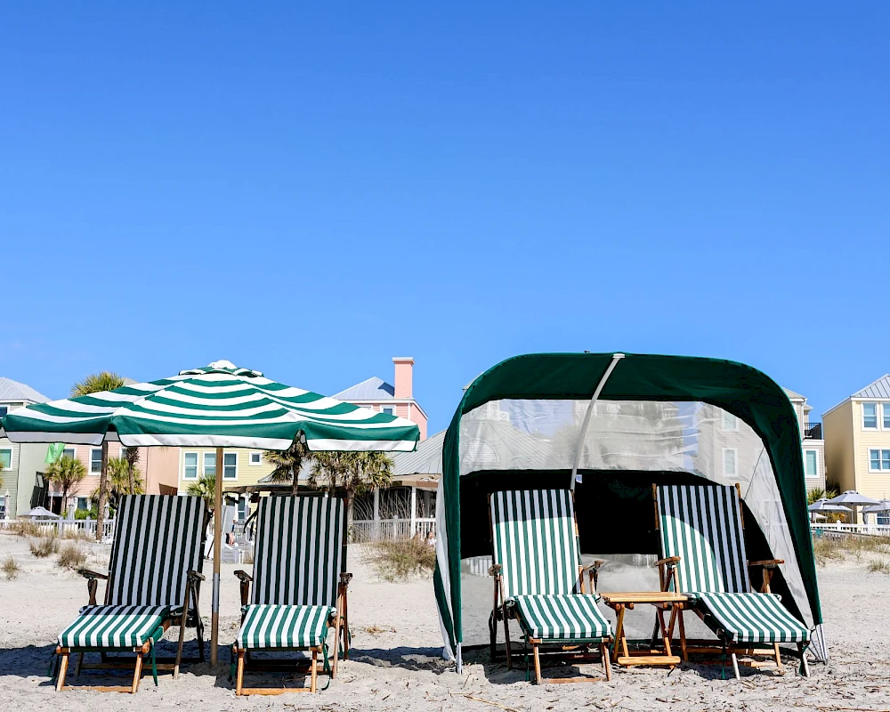 People relaxing on striped beach chairs under umbrellas and a canopy on a sandy beach with blue sky and seaside houses in the background.