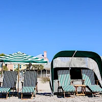 People relaxing on striped beach chairs under umbrellas and a canopy on a sandy beach with blue sky and seaside houses in the background.