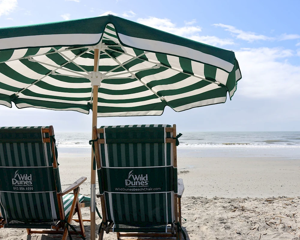 Two striped beach chairs beneath a green-and-white umbrella face the calm sea and sandy shore.
