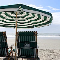 Two striped beach chairs beneath a green-and-white umbrella face the calm sea and sandy shore.