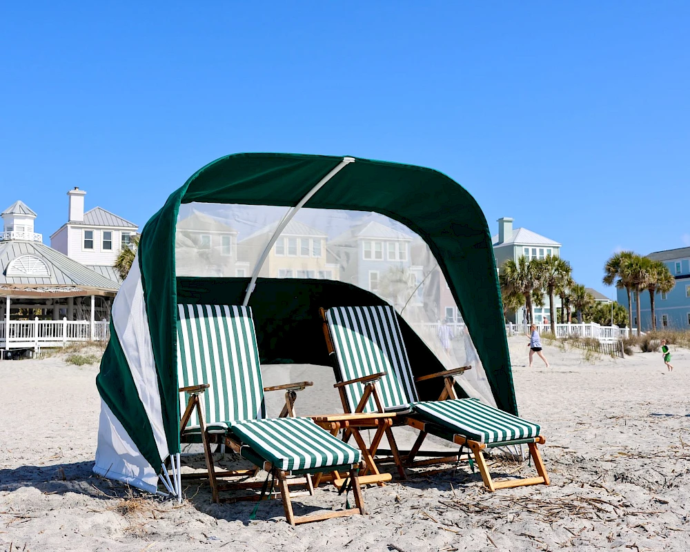A beach scene with a green-and-white cabana, four striped lounge chairs, and sand, with pale houses and palm trees in the background, under a clear blue sky.