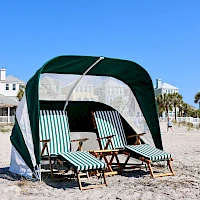 A beach scene with a green-and-white cabana, four striped lounge chairs, and sand, with pale houses and palm trees in the background, under a clear blue sky.