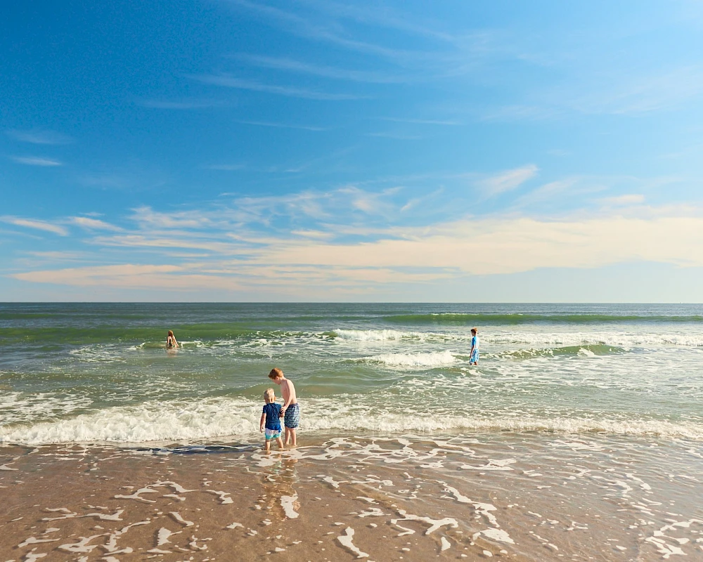 A sunny beach with blue skies, gentle waves, and a few people swimming and wading near the shore.