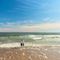 A sunny beach with blue skies, gentle waves, and a few people swimming and wading near the shore.