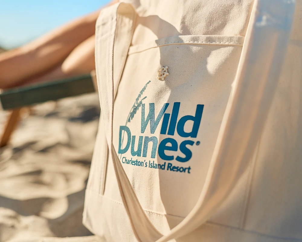 A sandy tote bag with the “Wild Dunes Charlestons Island Resort” logo on a beach, sunlit dunes in the background, outdoorsy vibes.