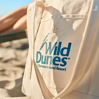 A sandy tote bag with the “Wild Dunes Charlestons Island Resort” logo on a beach, sunlit dunes in the background, outdoorsy vibes.