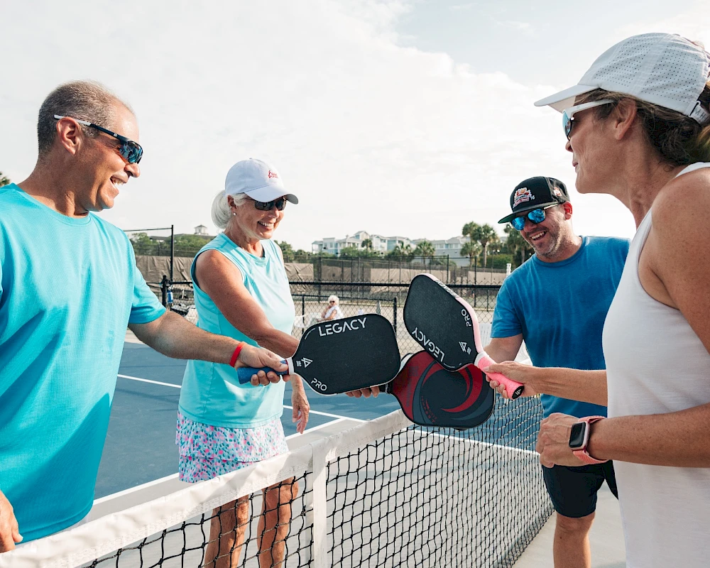 Four people on a pickleball court high-fiving over the net, prepping paddles and smiling as they chat near the fence.