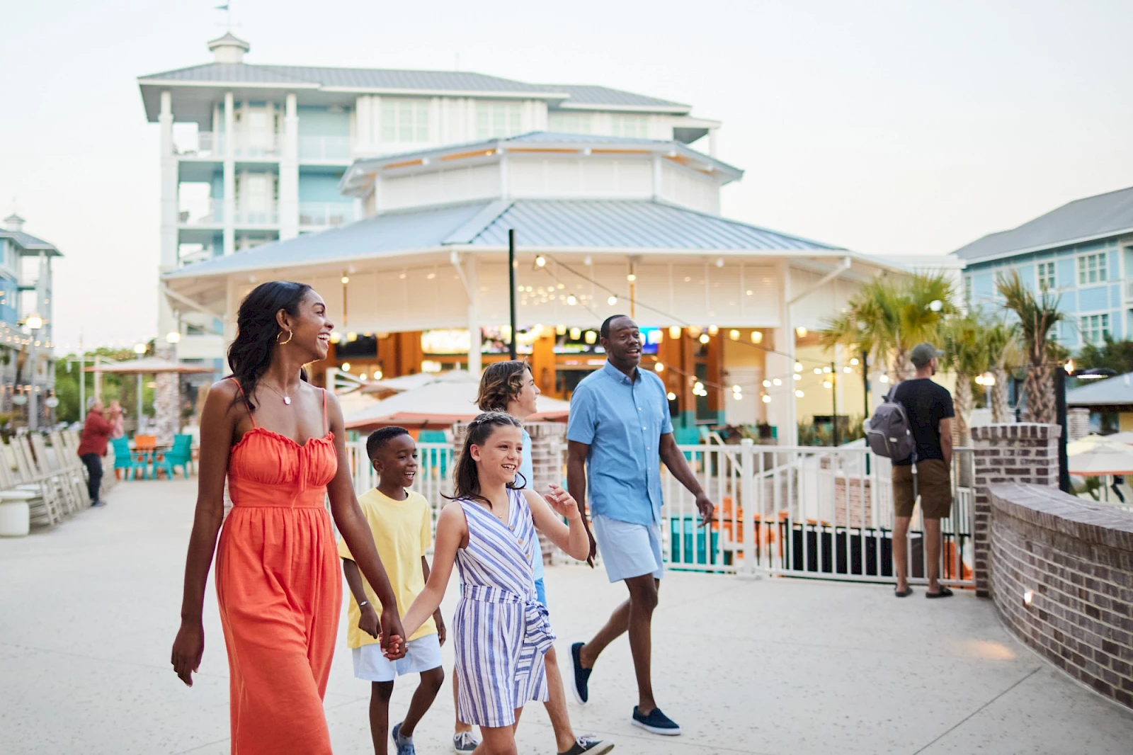 A family walks along a boardwalk by a resort, smiling and holding hands, with a white building and string lights in the background.