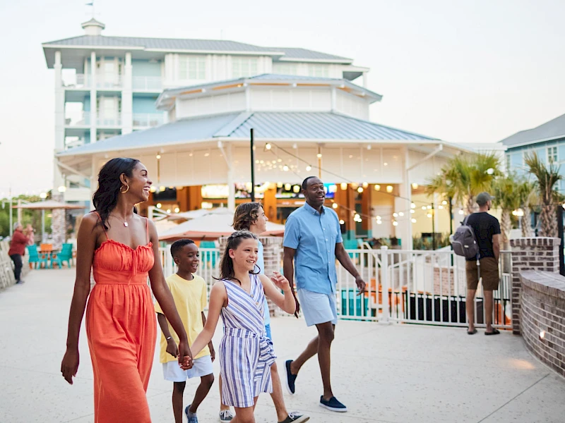 A family walks along a boardwalk by a resort, smiling and holding hands, with a white building and string lights in the background.
