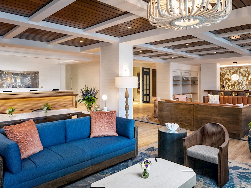 A stylish hotel lobby or lounge with a blue sofa, pink throw pillows, a white coffee table, and modern seating areas; warm lighting and chandeliers.