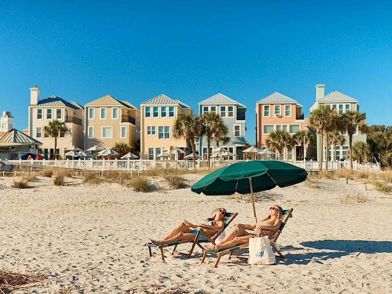 Three people lounge under a green beach umbrella on a sunny sandy beach, with colorful coastal houses in the background.