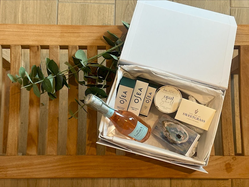 A white gift box open on a wooden bench contains skincare and beauty items, including bottles, jars, and a small card, with greenery beside it.