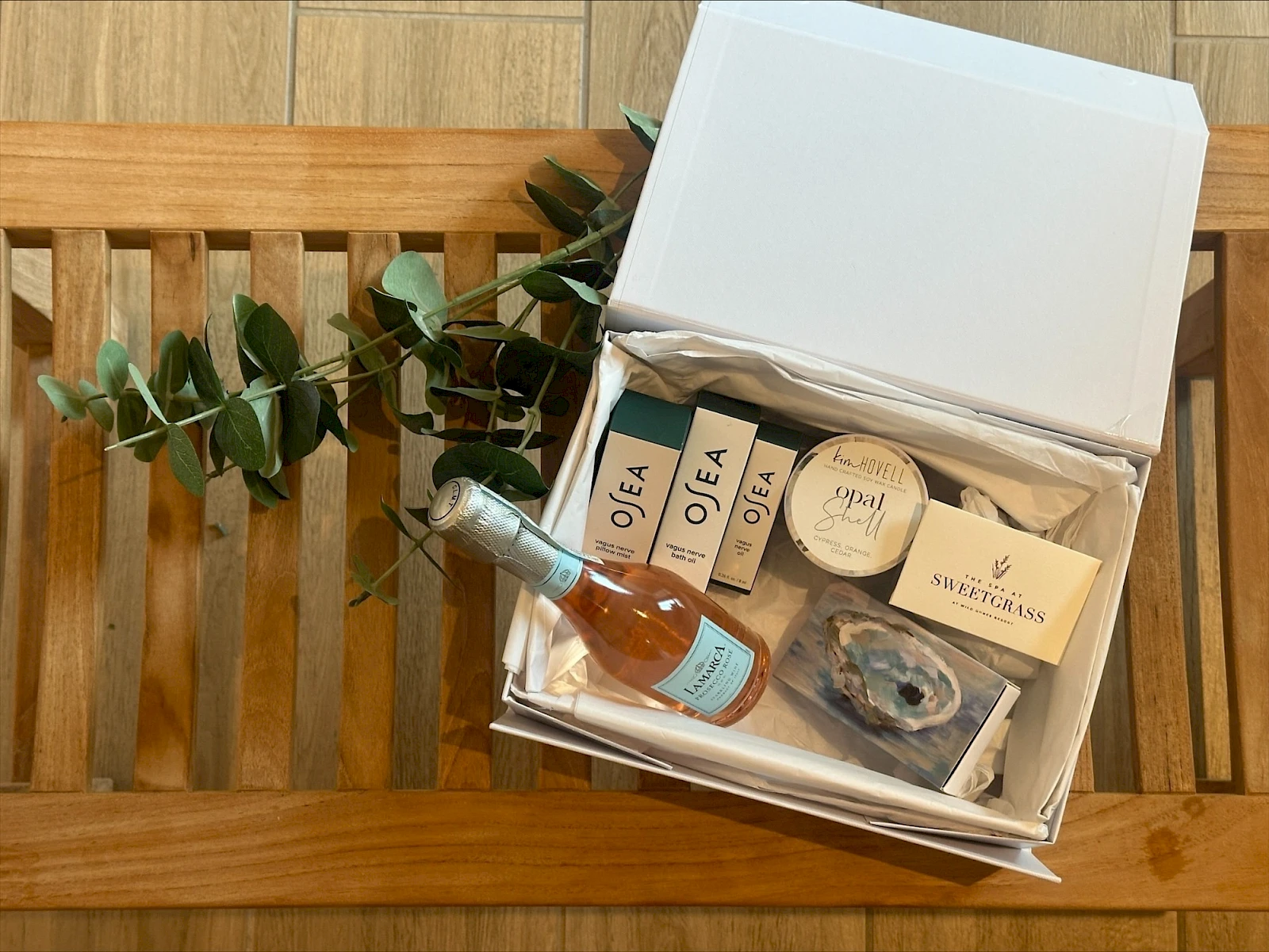 A gift box open on a wooden chair, containing a small bottle of rosé, soaps, a candle, and skincare items with greenery.