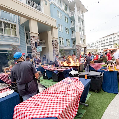 A lively outdoor barbecue party on a patio with multiple grills, people cooking and dining beside checkered red-and-white tables, in a residential complex.