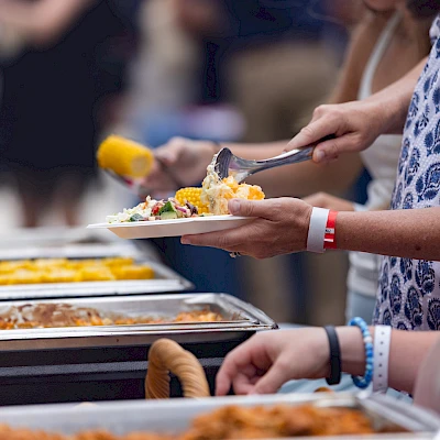 A lively outdoor barbecue party on a patio with multiple grills, people cooking and dining beside checkered red-and-white tables, in a residential complex.