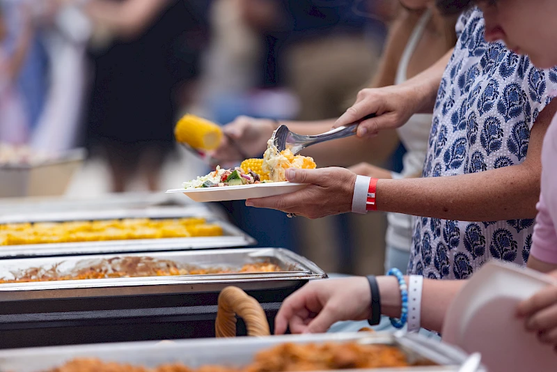 A lively outdoor barbecue party on a patio with multiple grills, people cooking and dining beside checkered red-and-white tables, in a residential complex.