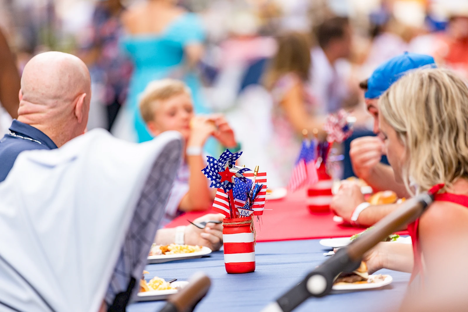 A long table outdoors with people eating, decorated with small American flags and red-white-blue cups, under a festive patriotic setting.