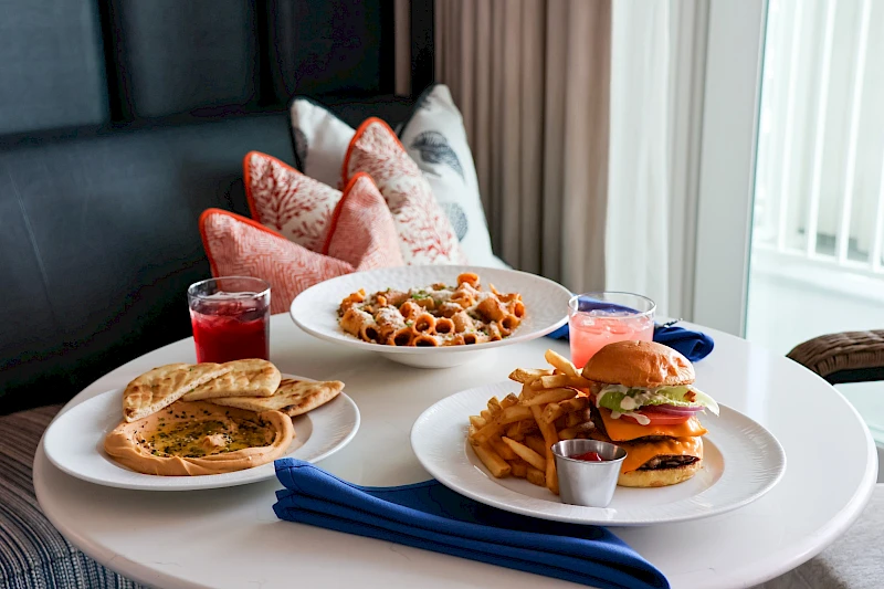 A cozy table with multiple dishes: quesadilla/pita-style bread, a burger with fries, a pizza-like flatbread, and a pasta dish, all served with sauces in small cups, on a white round table by a window, top it at 140 characters, always ending the sentence.