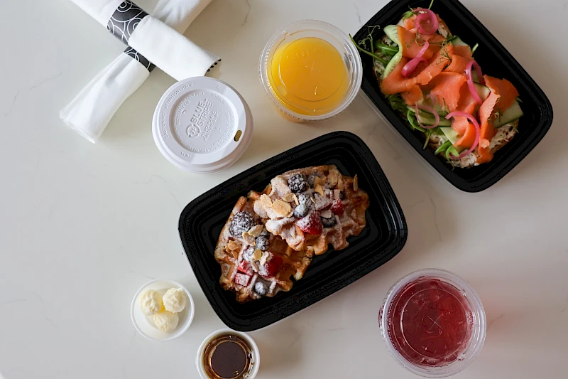 A top-down view of a meal: two black takeout containers with a chicken dish and a salad, plus a small cup of butter, orange juice, a red beverage, and utensils on a white table.