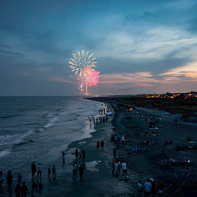 Fireworks light up the twilight sky over a crowded beach, with people gathered along the shoreline watching the display.